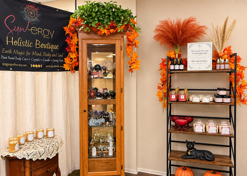 A styled boutique interior corner with a tall wooden glass-front cabinet, a black metal shelf with candles, jars, decorative orange leaves, pumpkins, and dried grasses, plus a sign and wall banner reading Sunnygro Holistic Boutique.