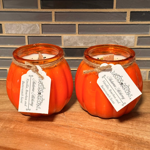 Two orange pumpkin-shaped candles with tags on a wooden surface and tiled wall background.