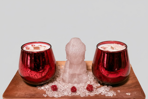 Two red candle holders with Rose Quartz Buddha on a wooden surface, surrounded by rose quartz stones and small red flowers, against a white backgound.
