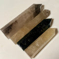 Four polished crystal points arranged diagonally on a plain light background, including smoky gray, tan, black, and pale yellow varieties.