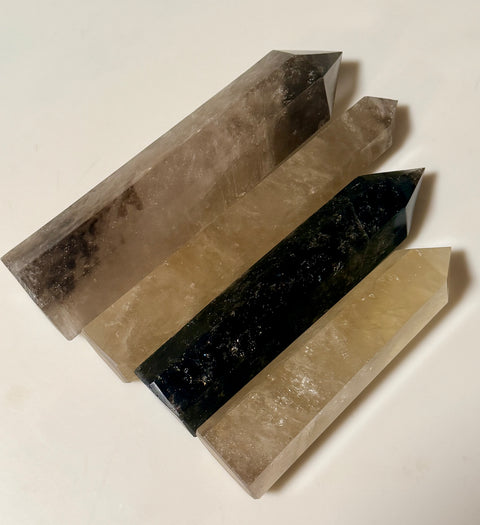Four polished crystal points arranged diagonally on a plain light background, including smoky gray, tan, black, and pale yellow varieties.