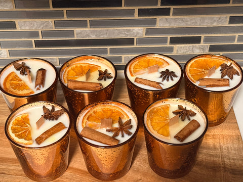 Seven bronze candle holders with soy candles topped with orange slices, cinnamon sticks, and star anise on a wooden surface.