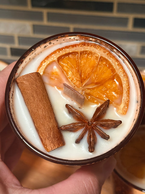 Close-up of a soy candle with orange slices, cinnamon stick, and star anise against a brick wall.