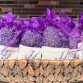 Lavender sachets filled with dried lavender buds, tied with purple organza ribbons, and arranged in a woven basket with white paper bands printed with a purple botanical design.
