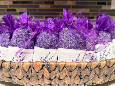 Lavender sachets filled with dried lavender buds, tied with purple organza ribbons, and arranged in a woven basket with white paper bands printed with a purple botanical design.