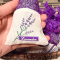 A hand holding a small lavender sachet made of cream fabric with a purple embroidered lavender stem and a purple label reading 'Lavender.' More purple lavender sachets are visible in a woven basket behind it against a tiled wall background.