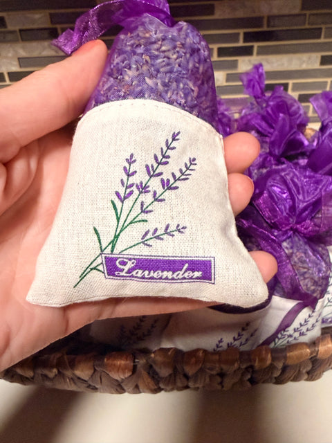 A hand holding a small lavender sachet made of cream fabric with a purple embroidered lavender stem and a purple label reading 'Lavender.' More purple lavender sachets are visible in a woven basket behind it against a tiled wall background.