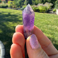 Hand holding amethyst crystal pendant outdoors with greenery in the background