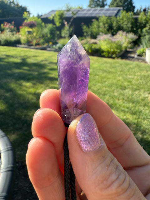 Hand holding amethyst crystal pendant outdoors with greenery in the background